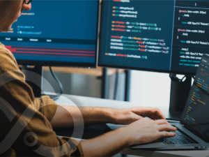 A person typing on a laptop at a desk with three monitors displaying colorful programming code.