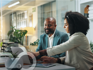 Two coworkers collaborate in an office, reviewing information on a laptop as one points to the screen.