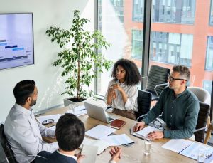 Responsible AI - three business men and a business woman sit in a meeting room with laptops on the desk, a plant in the background